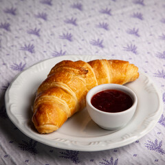 A delicious french traditional croissant bread next to a little cup of grape jelly on a white plate above a delicate tablecloth in a house kitchen or restaurant ready for breakfast or afternoon snack