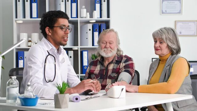 care health, black doctor will measure pressure with modern tonometer to an elderly man at a reception with his wife in medical office
