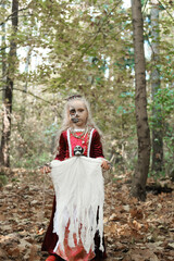 A child girl poses in a forest in a medieval dress in the image of a dead princess with makeup on her face. Girl holding a skeleton doll