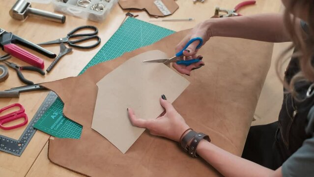High angle medium close up of hands of woman cutting out cardboard pieces to make leather bag for client in workshop