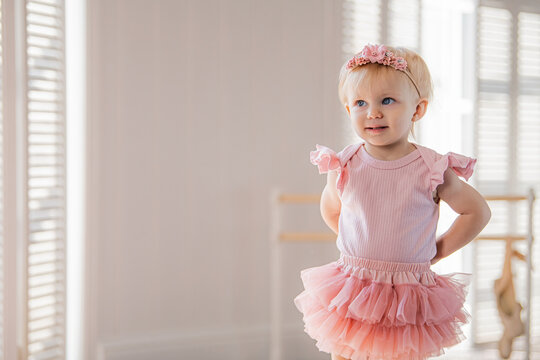 A cute little ballerina in a pink ballet costume is standing near the ballet barre in the room. Kid and ballet dance. Copyspace.