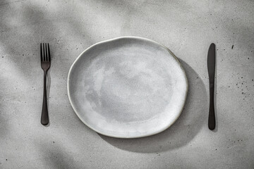 Black fork and knife near gray plate placed on concrete texture. Minimal table setting as...