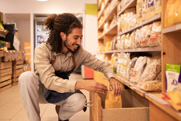 Smiling young ethnic male seller picking goods from shelf in store