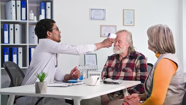 health care, professional black male nurse examines an old man with his wife and measures temperature with a non-contact thermometer in medical office