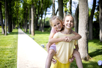 Two girls sisters of school age hug each other. They laugh and fool around. A walk through the summer city park. Love and friendship within the family