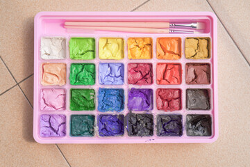 Dried colorful gouache in pink painting palette box with two pieces of white paint brush. Cracked water color and small brush.