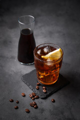 Cold brew coffee in a glass with ice and lemon on a dark concrete background with coffee beans and bottle. Concept summer craft refreshing homemade drink.