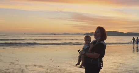 happy family Asian mother carrying cute baby daughter walking on tropical beach during amazing sunset sky enjoying their vacation together at ao nang beach, krabi Thailand