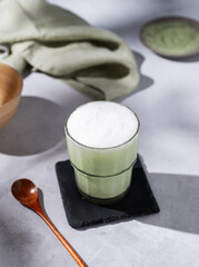 Matcha latte with ice. Green tea with milk in a glass on a light background with spoon, napkin and morning shadows. The concept of a traditional healthy Japanese summer refreshing drink.