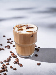 Iced coffee latte in a glass with milk on a light background with coffee beans and morning shadows. Summer refreshment concept.