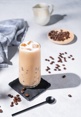 Iced coffee latte in a tall glass with milk on a light background with coffee beans, milk jug and morning shadows. Summer refreshment concept