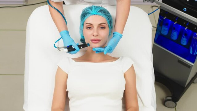 View of doctor cosmetologist doing anti aging procedure in cosmetology office. Satisfied woman in disposable hat lying on couch and relaxing. Working with Apparatus.