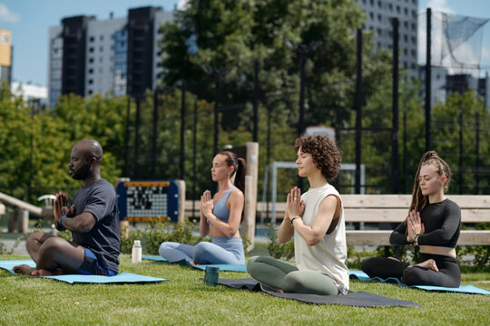 Side view of group of intercultural athletes practicing yoga outdoor while sitting on mats with their legs crossed and palms put together by chest - Powered by Adobe