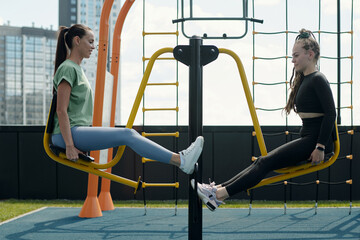 Side view of two young modern sportswomen in activewear sitting on sports facilities in front of camera and doing physical exercises