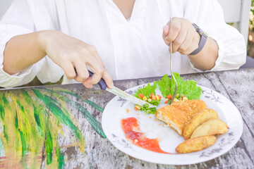 Obraz na płótnie Canvas A woman wearing white clothes is slicing steak on a ceramic plate on a wooden table. Fried chicken steak served with tomato sauce and mixed vegetables. Concept for healthy food, fat loss, weight loss.