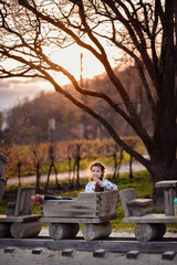 Little schooler girl with longhair on spring sunset sitting in wooden train on playground. Big tree behind her