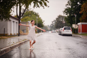 Little preschooler girl in white dress with black hearts dancing and spinning on wet empty street under the rain
