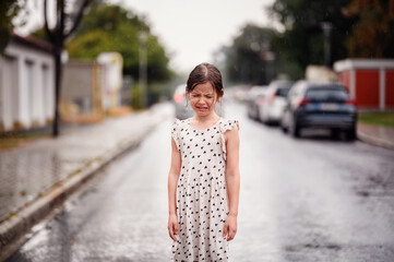 Little preschooler girl in white dress with black hearts standing alone on wet empty street under...