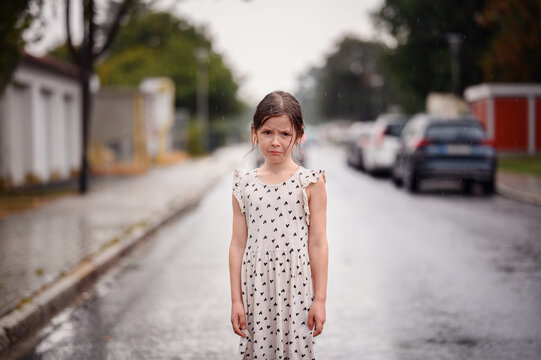 Little preschooler girl in white dress with black hearts standing alone on wet empty street under the rain. Close-up portrait