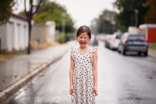 Little preschooler girl in white dress with black hearts standing alone on wet empty street under the rain. Close-up portrait
