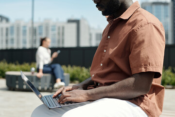 Cropped shot of young African American male analyst pressing keys of laptop keyboard while sitting in front of camera in urban environment