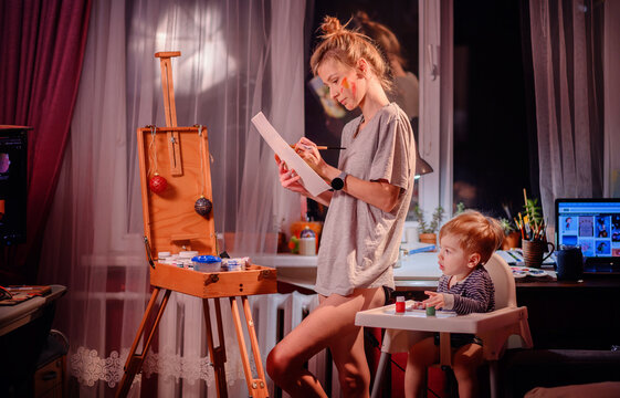 Young blonde woman drawing a picture in hands while little toddler son playing with jars of paint