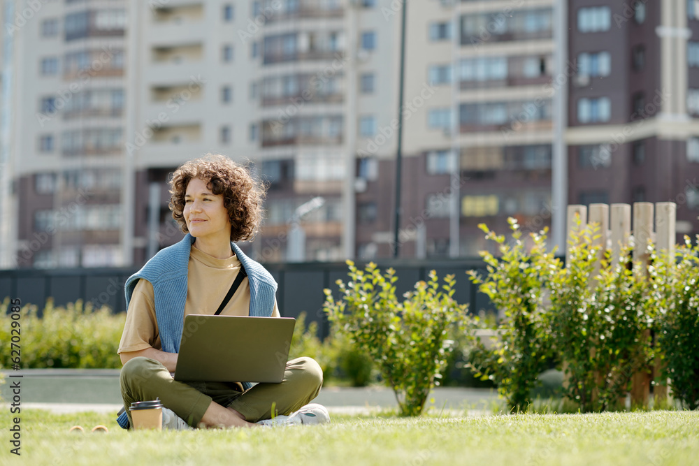 Poster young smiling woman with laptop sitting on green grass in park on sunny summer day and looking aside