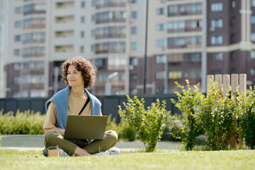 Young smiling woman with laptop sitting on green grass in park on sunny summer day and looking...
