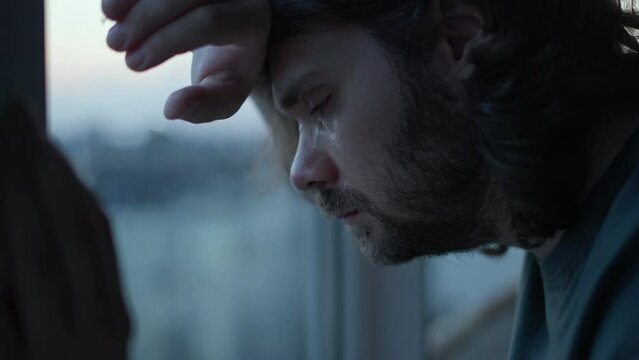 Depressed man crying, thinking about problems near window. Close up of pensive, sad depressed unhappy beard man with long hair. Mental health, man in tears, man cry depression concept. Authentic