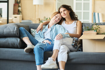 Two happy young girlfriends in casualwear sitting on comfortable couch in living room and enjoying rest after relocation to new apartment