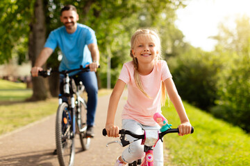 Day in the park. Happy family of caucasian father and girl riding bicycles outdoors in the summer, smiling at camera