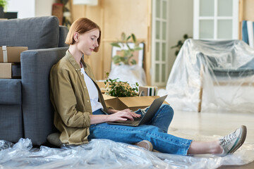 Side view of young female solopreneur in casualwear sitting on the floor by couch in living room and typing on laptop keyboard while networking