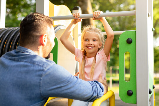 Tales from the playground. Father playing with his little happy daughter in the park, girl sitting on slide