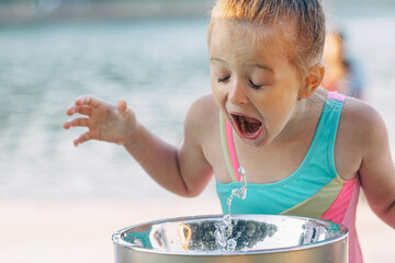 A little blonde girl drinks from a drinking fountain in a city park on a hot summer day outdoors.