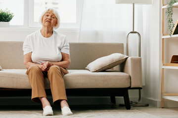 Elderly woman severe abdominal pain sitting on the sofa, health problems in old age, poor quality of life. Grandmother with gray hair holding her stomach, poisoning, problems with stool.