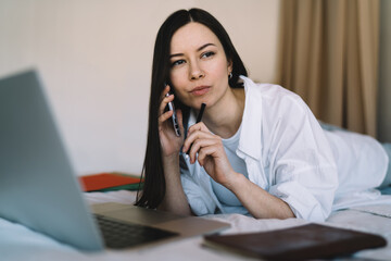 Pensive woman speaking on smartphone during remote job via computer