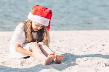 Happy little girl in Santa Claus hat has fun on a sandy beach. Little girl celebrates Christmas in...