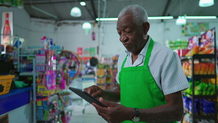 African American senior employee of supermarket using tablet device standing inside grocery store,...