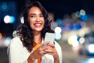 Portrait of happy young woman using smartphone standing on the night city street full of neon...