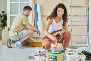 Young brunette woman in coveralls mixing beige liquid paint in plastic bucket while sitting on squats in front of camera against her husband