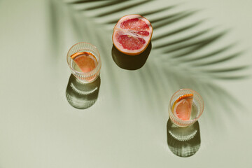 Summer flat lay with two glasses of refreshment drinks and grapefruit slices on green background with palm leaf shadow sun and sunlight. Vacation, holiday, mocktail, creative minimal concept.