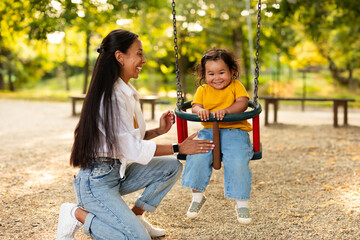 Obraz premium Glad Millennial Asian Mother Swinging Baby Daughter At Playground Outside