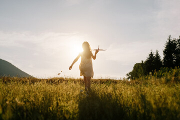 Silhouette of playful preteen girl kid run with airplane on background amazing summer warm sunset and majestic mountains. Dream freedom concept. Child runs on field holding in hands toy aircraft