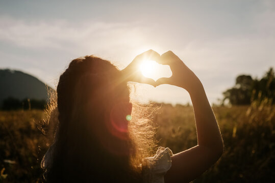 Back view of little girl child making heart with hands during calm sunset or sunrise at warm summer day. Sunbeams Sunlight. Happy Valentines day, Mothers Day, love nature or life happiness concept