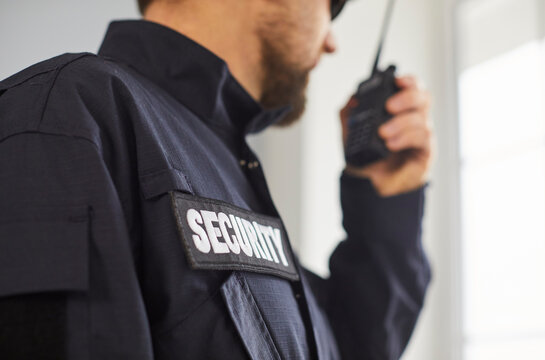 Close up portrait of confident male security guard standing by the window. Guardian man in uniform talking by the walkie talkie standing indoors and looking to the side. Selective focus.