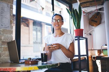 Portrait of successful female copywriter with laptop and modern cellular gadget smiling at camera...