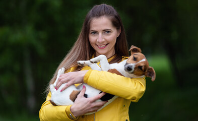 Young woman in yellow jacket holding her Jack Russell terrier dog on hands, blurred green trees background