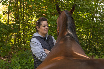 50-jährige Pferdezüchterin mit schwarzen Haaren steht nachdenklich neben ihrer hochtragenden braunen Stute im Wald