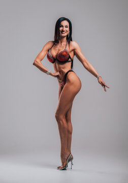 Smiling athletic woman in red bikini showing muscles on gray background.
