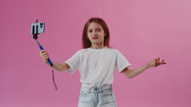 Young girl with a selfie stick making a funny video on a smartphone on a pink background. Concept of the child's desire to be a famous creative blogger